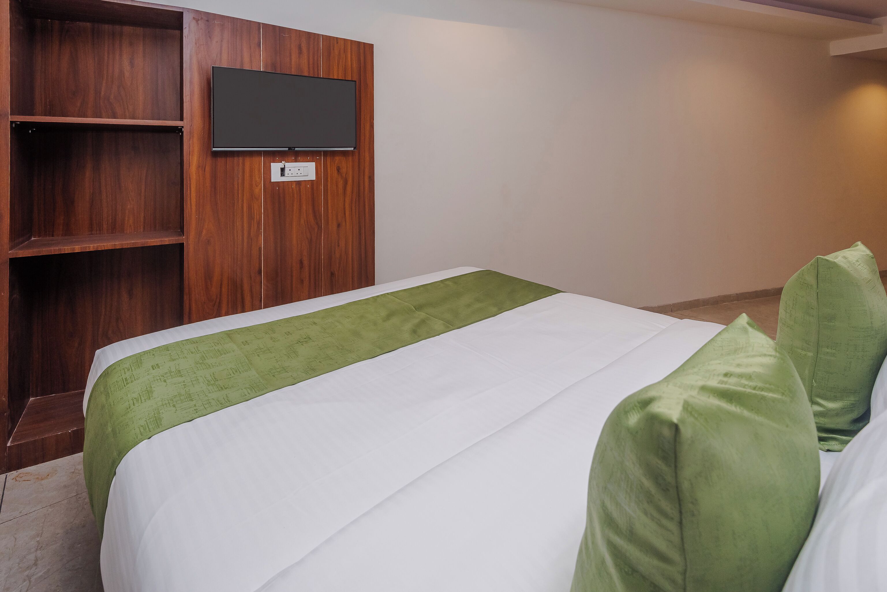Una habitación de hotel con una cama, un edredón verde, un televisor fijado a un panel de madera y una pared blanca.