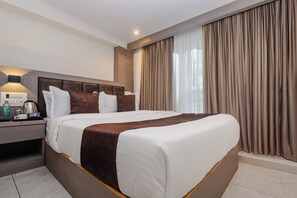 A hotel room with a large bed, a bedside table with a lamp, a window with curtains, and a wall-mounted thermostat.