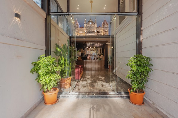 Entrada de hotel con puerta de vidrio, plantas en macetas y vista de un edificio con torre de reloj.