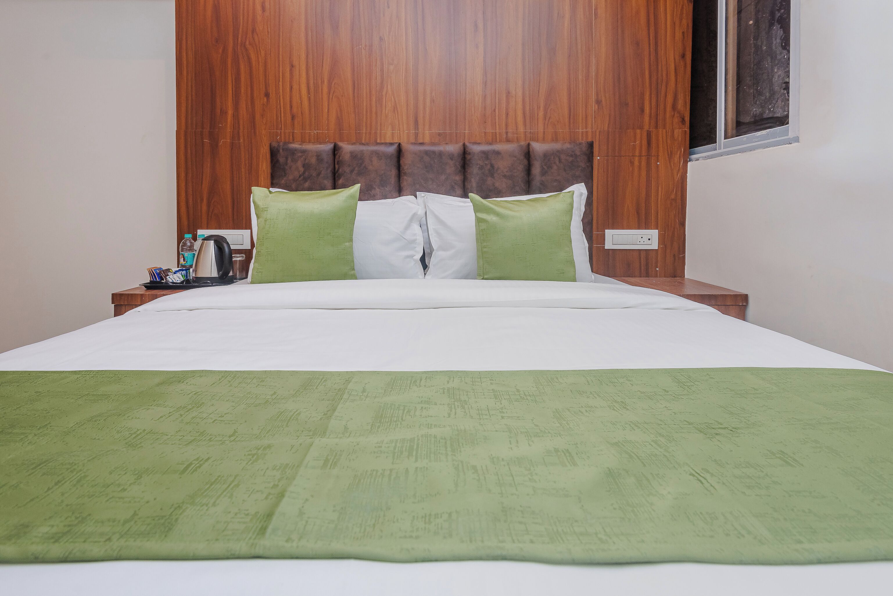 Une chambre d’hôtel avec une tête de lit en bois, un lit avec une literie blanche et verte, un plaid vert et une table de chevet avec une bouilloire et une tasse.