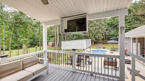 Una terraza techada con televisor, asientos exteriores y vistas a un jardín con piscina y mobiliario de patio.