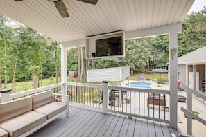 Eine überdachte Terrasse mit Fernseher, Sitzgelegenheiten im Freien und Blick auf einen Garten mit Pool und Gartenmöbeln.