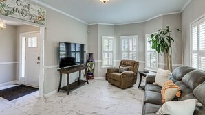 Una sala de estar con televisor, sofá, sillón, planta y un letrero decorativo.