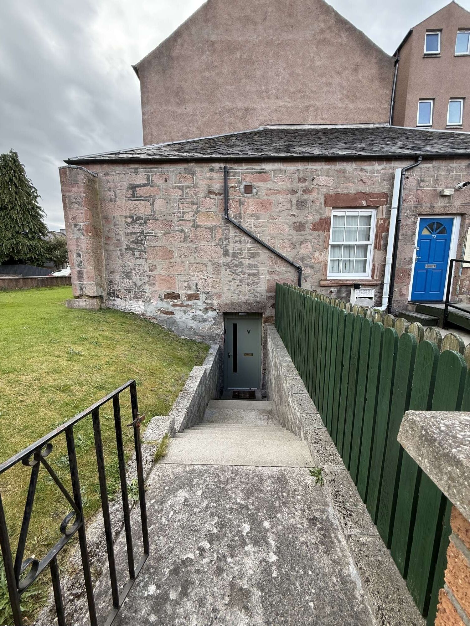A stone building with a narrow entrance, a blue door, and a green fence.