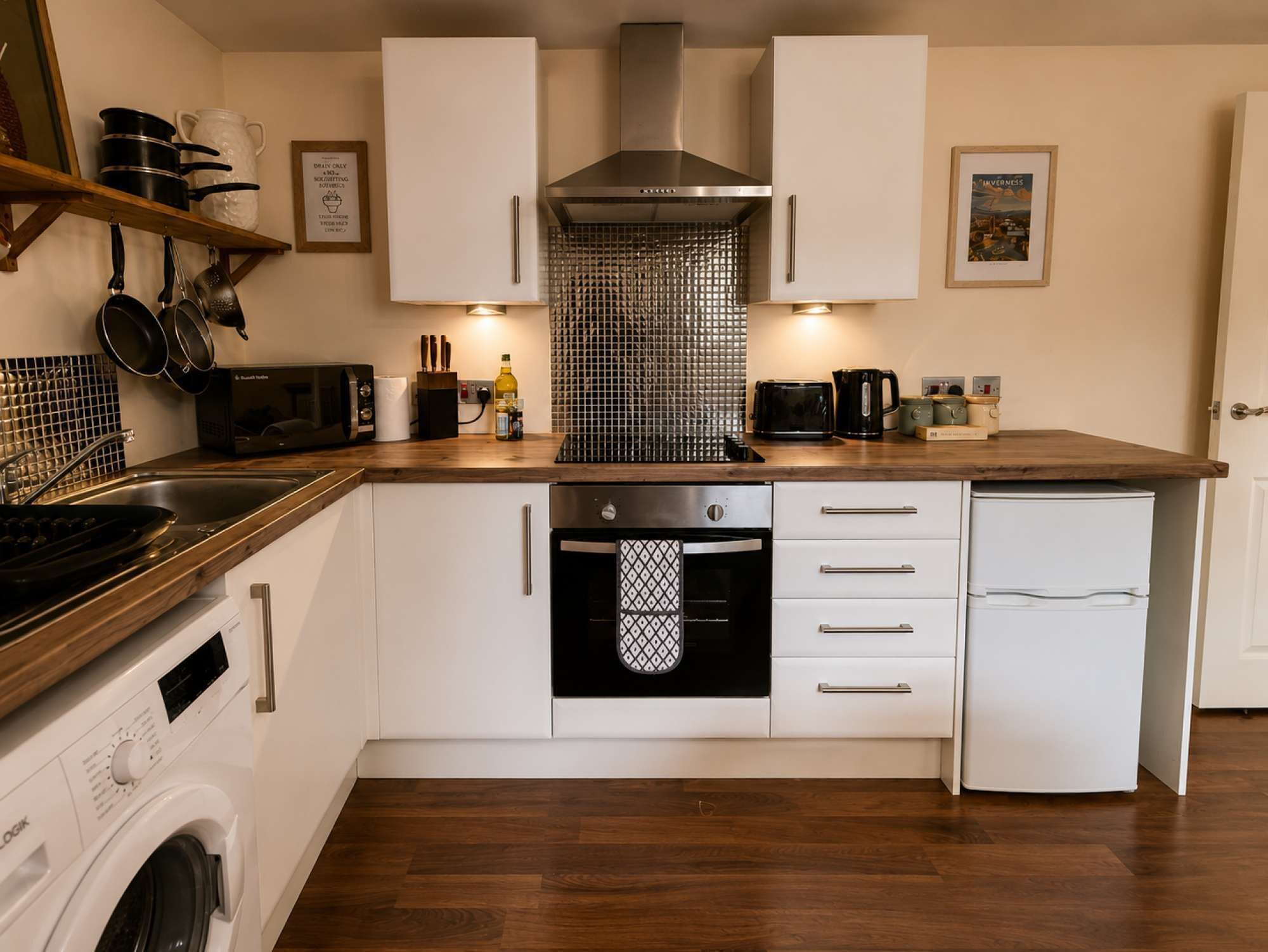 A compact kitchen with white cabinets, a stainless steel range hood, a built-in oven, a dishwasher, and a washing machine.