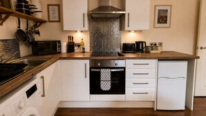 A compact kitchen with white cabinets, a stainless steel range hood, a built-in oven, a dishwasher, and a washing machine. - Heather & Stone Bothy, Inverness (Inverness)