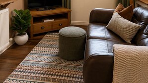 A living room with a leather sofa, ottoman, TV on a wooden stand, a potted plant, and a window with curtains. - Heather & Stone Bothy, Inverness (Inverness)
