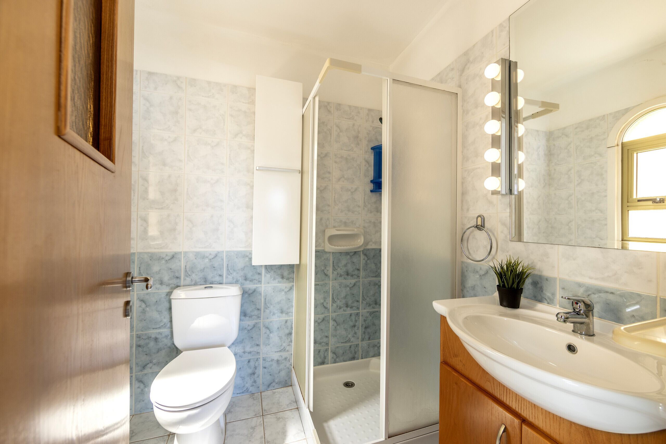 A bathroom with a toilet, shower, sink, and mirror with lights.