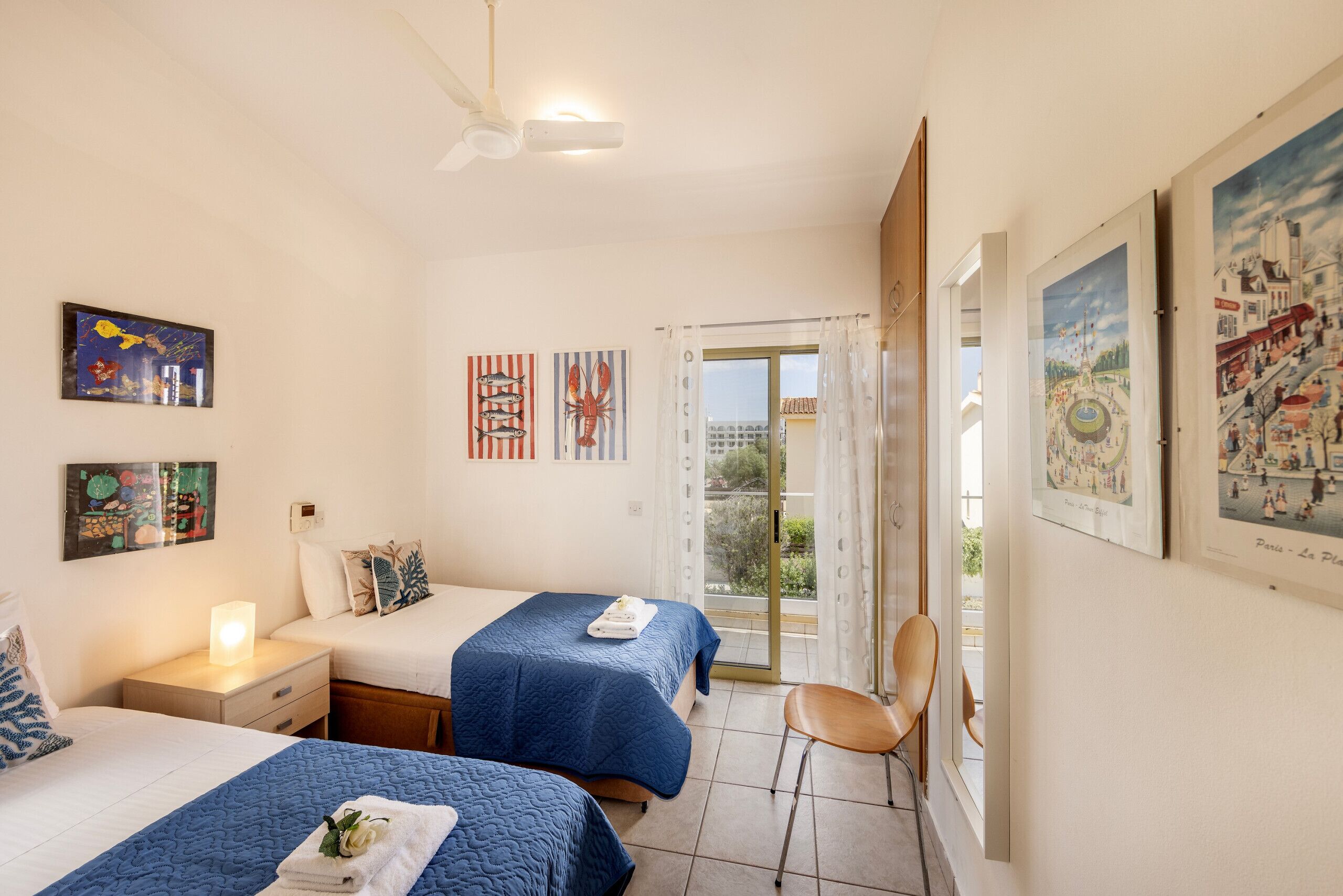 Camera d'albergo con due letti, una ventola a soffitto, opere d'arte alle pareti e vista attraverso il portavetro.