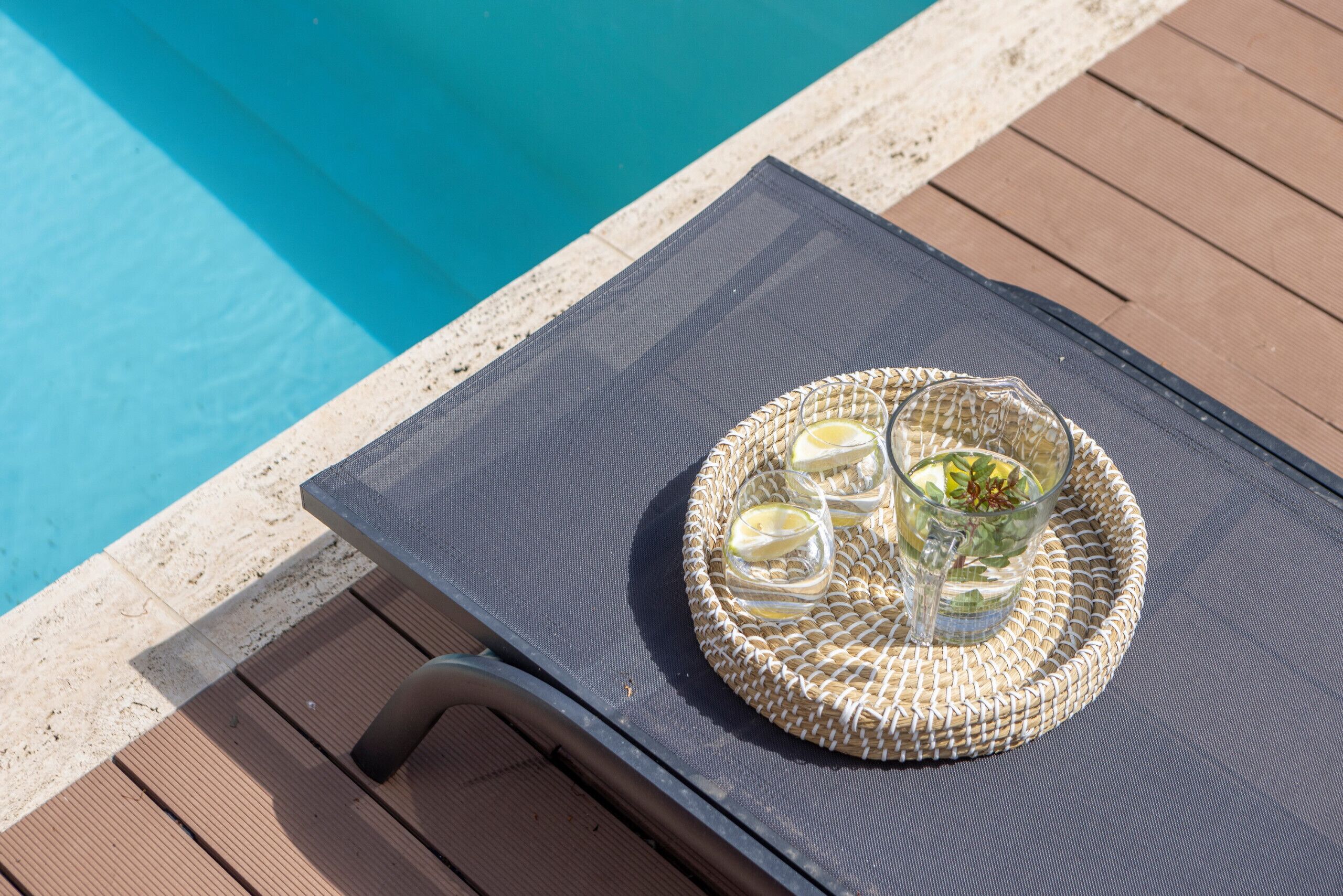 A poolside lounge chair with a woven tray table holding glasses and a beverage.