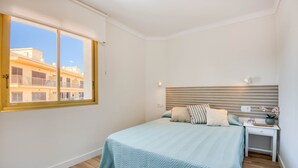 A bedroom with a large window, a bed with a striped headboard, a bedside table with a lamp, and a view of buildings outside. - RIELLS: Fully renovated seafront apartment (L'Escala)