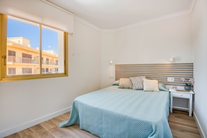 Ein Schlafzimmer mit großem Fenster, einem Bett mit gestreifter Kopfteil, einem Nachttisch mit Lampe und Blick auf Gebäude.
