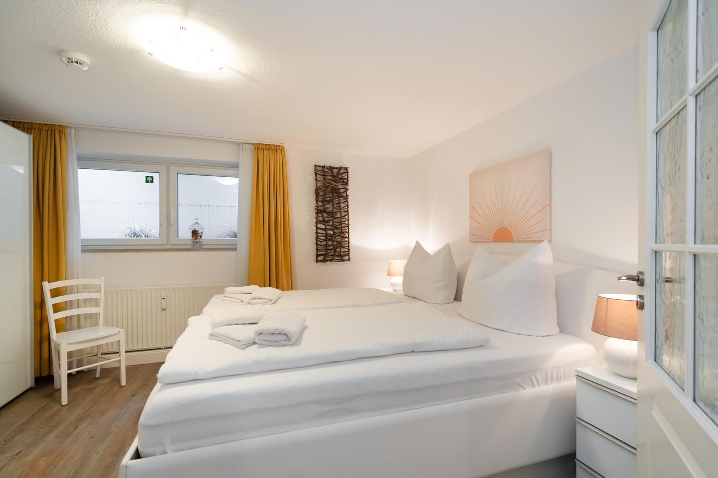 Ein Hotelzimmer mit Bett, Sessel, Fenster mit Vorhängen, Heizkörper und einem Wandbild.
