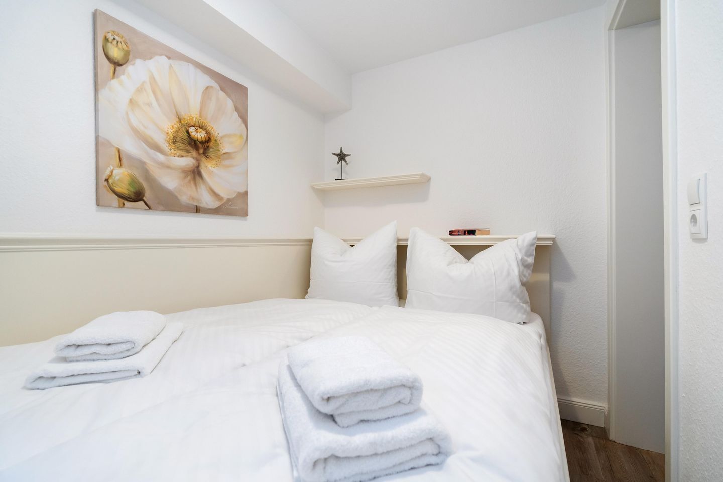 A neatly made bed with white linens and folded towels, a wall-mounted shelf with a book, and a large floral painting above the bed.