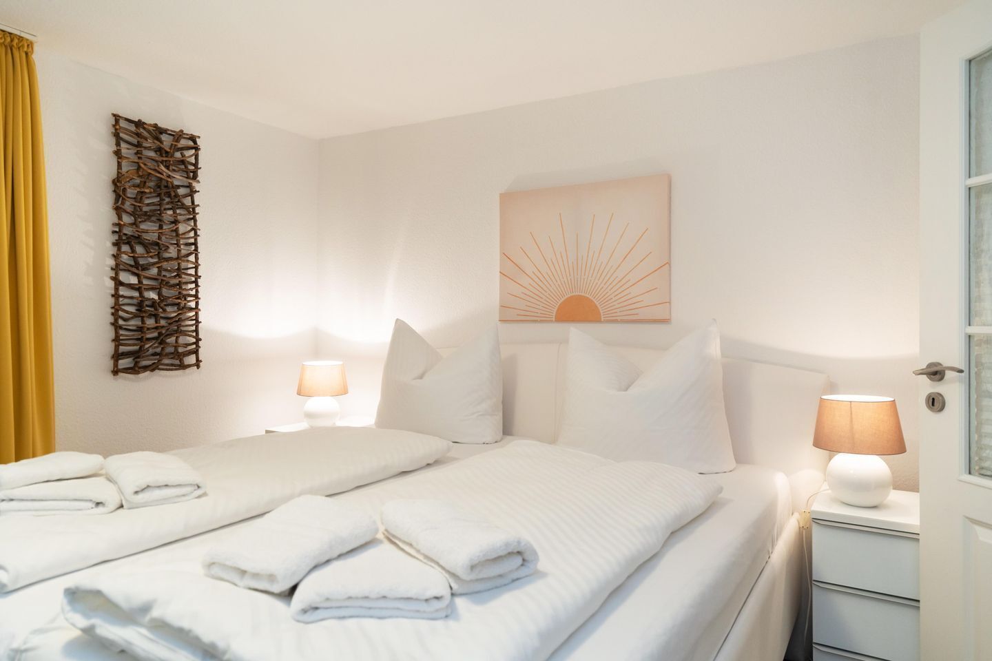 Hotellihuone, jossa on kaksi sänkyä, valkoiset vuodevaatteet, aurinkoteemainen taideteos, seinälle asennettu abstrakti veistos ja yöpöytälamput.
