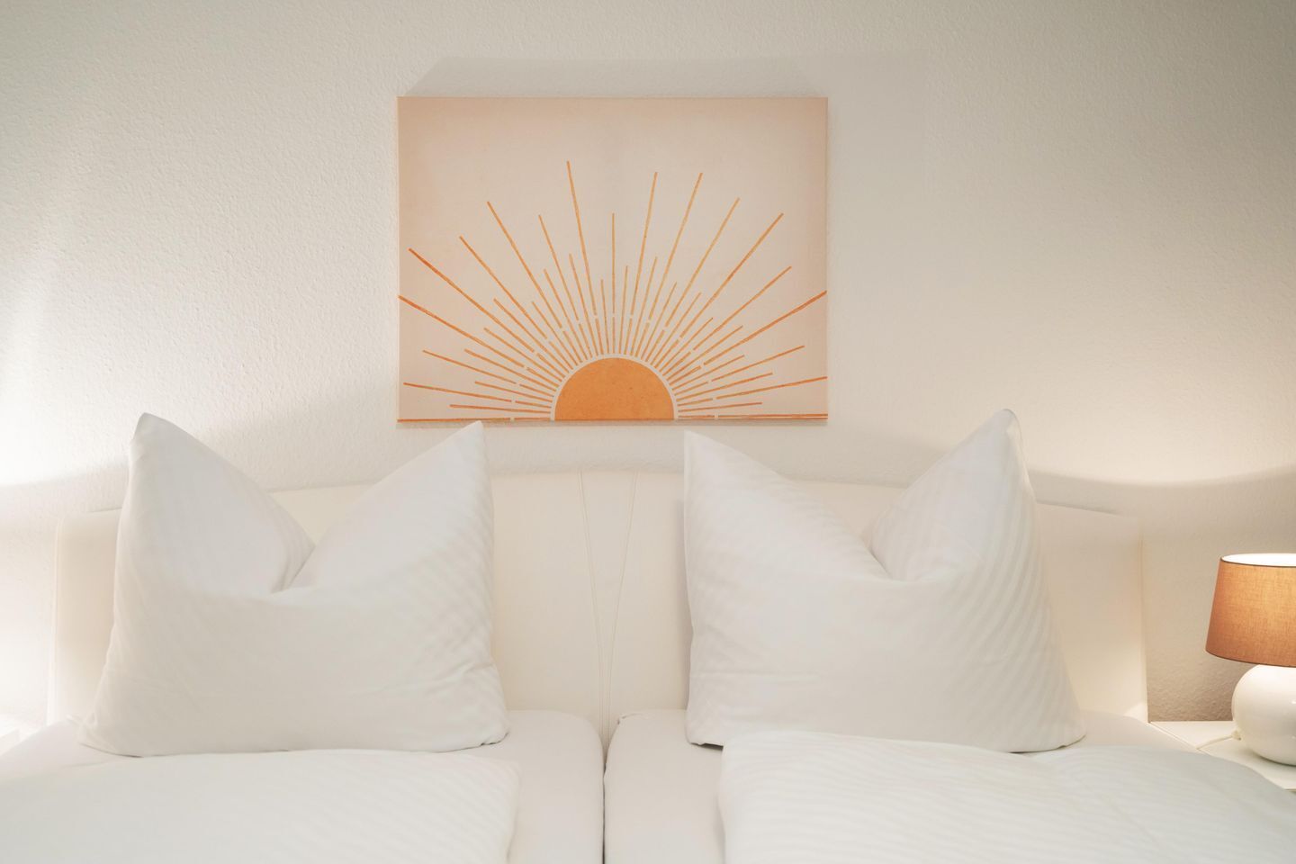 Ein Hotelzimmer mit Bett, weißer Bettwäsche, einem Kopfteil und einem an der Wand hängenden Kunstwerk mit einer Sonne.