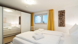 Een hotelkamer met twee bedden, een raam met gele gordijnen, een witte stoel en een decoratief wandobject.