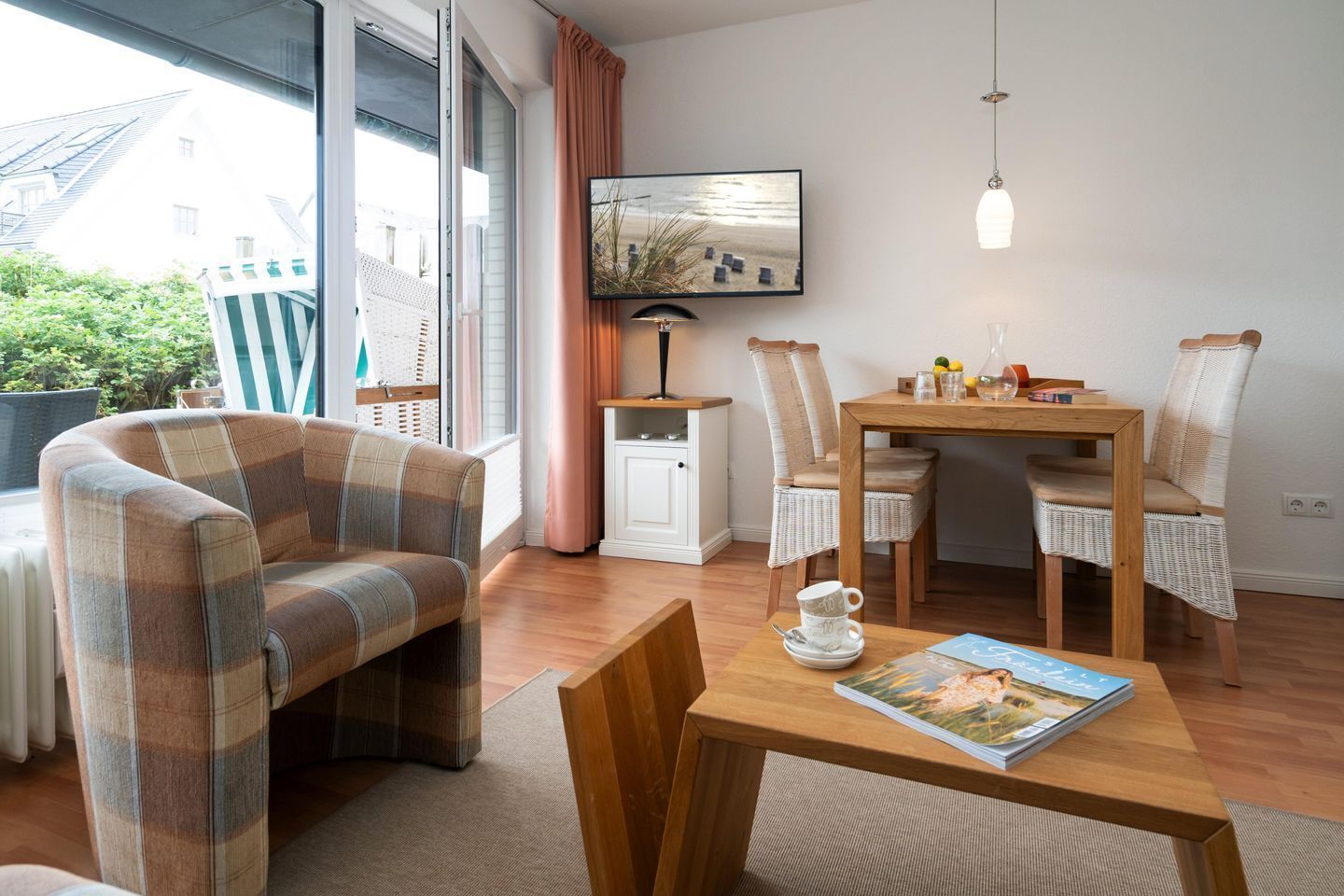 A cozy living room with a plaid sofa, a wooden dining table with chairs, a television mounted on the wall, and a window with curtains.