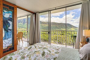 Hotelkamer met een groot bed, uitzicht op een bergachtig landschap door een glazen schuifdeur, een houten raam met glas-in-lood en een balkon met een stoel en tafel.