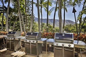 Drie roestvrijstalen Genesis gasgrills in een buitensetting met palmbomen en een bergachtige achtergrond.