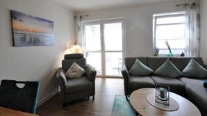 Ein Wohnzimmer mit Sofa, Sesseln und Couchtisch, einem Gemälde an der Wand und Blick durch eine Glastür.