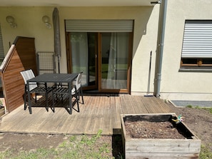 A patio with a table and chairs, a wooden deck, a sliding glass door, and a window with a shutter. - Saxon Lake Retreat in Birkwitz – Relax Near Saxon Switzerland (Pirna)