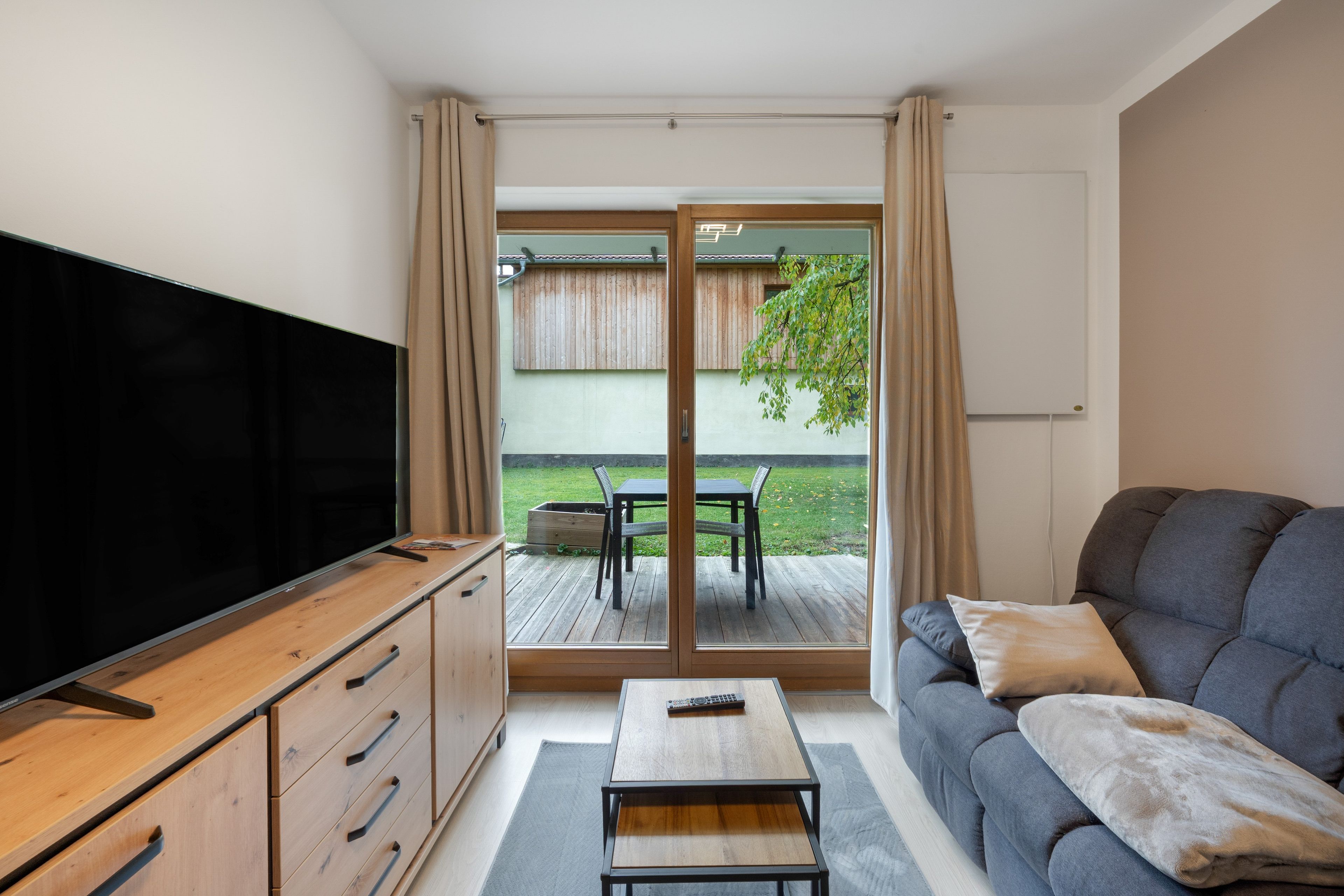 A living room with a flat-screen TV, a sofa, a coffee table, and a sliding glass door leading to a patio with a table and chairs.