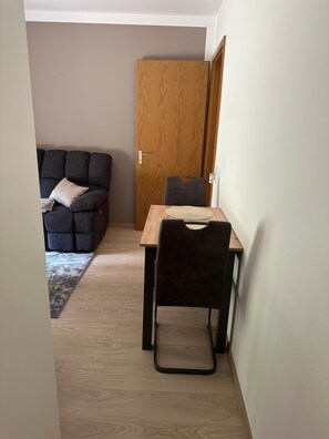 Ein Zimmer mit Holztür, einem dunklen Sofa mit Kissen, einem kleinen Tisch mit Stuhl und einem gemusterten Teppich auf dem Boden.