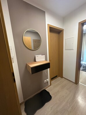 Un pasillo con una puerta de madera, un espejo en la pared, un estante blanco con un dispositivo y un tapete negro en el suelo.