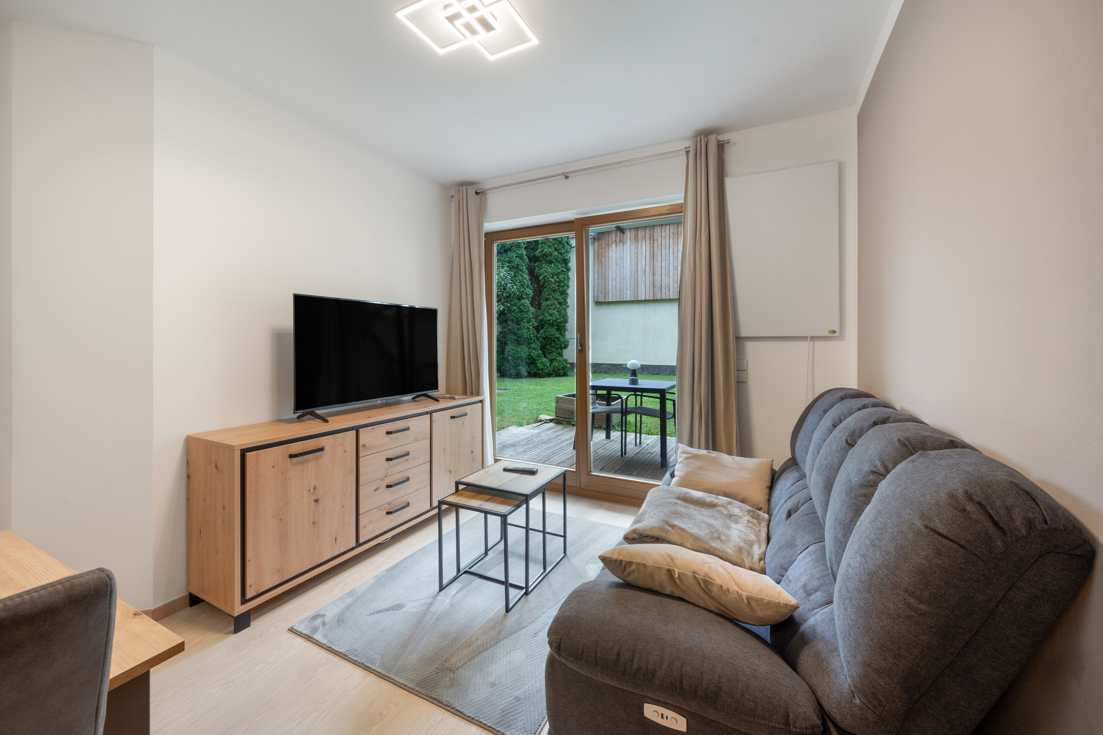 A modern living room with a flat-screen TV, wooden cabinet, grey sofa, and a small table.