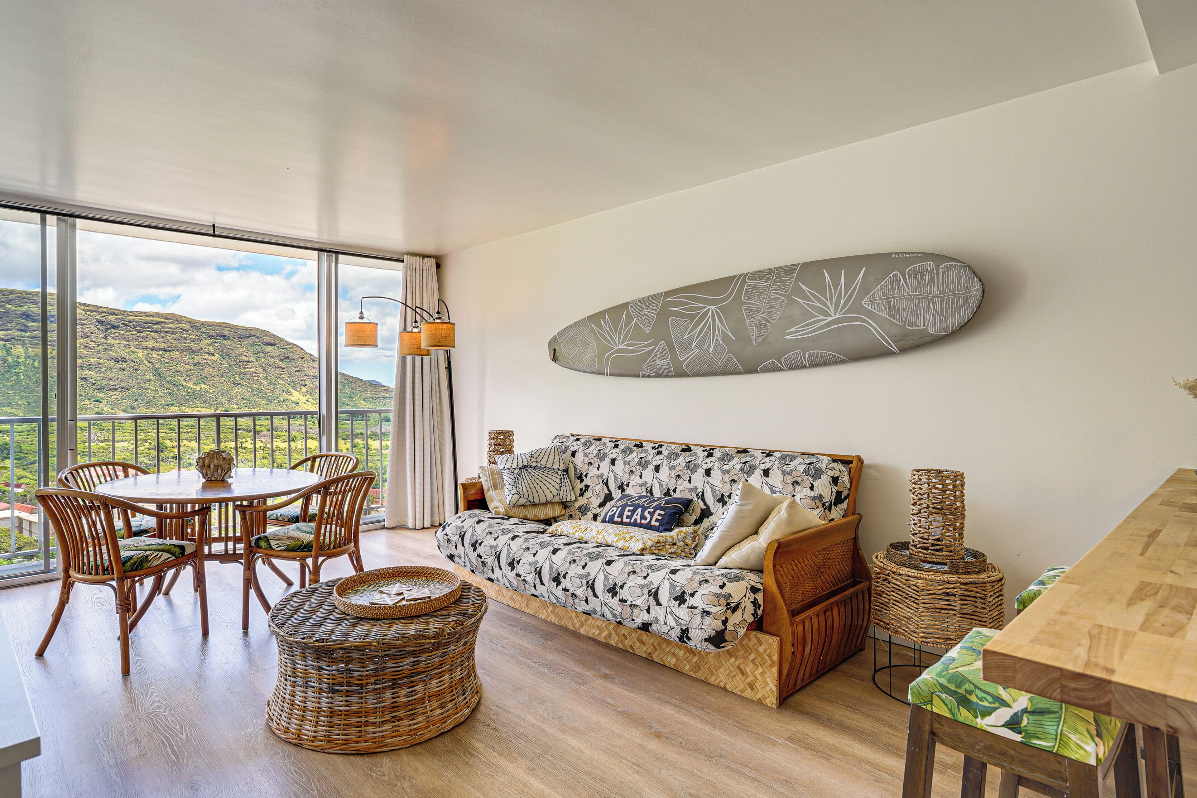 A room with a sofa, dining table, wicker chairs, and a view of a mountainous landscape.