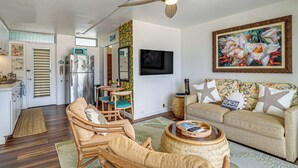 A living room with a ceiling fan, a television, a sofa, and a dining area with a refrigerator. - Steps to Beach & Pool! Oceanfront Waianae Studio (Waianae)