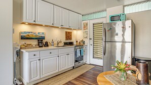 A kitchen with white cabinets, a stainless steel refrigerator, a microwave, and a wooden dining table with a vase of flowers. - Steps to Beach & Pool! Oceanfront Waianae Studio (Waianae)