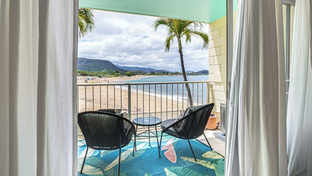 A balcony with two chairs and a table, overlooking a beach and palm trees. - Steps to Beach & Pool! Oceanfront Waianae Studio (Waianae)