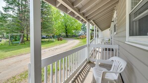 A covered porch with a white railing, a white plastic chair, and a view of a park with a swing set and trees. - Boat, Fish & Hike! All-Season Schroon Lake Getaway (Schroon Lake)