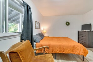 Un dormitorio con cama, una silla, cómoda, televisor y ventana con cortinas.