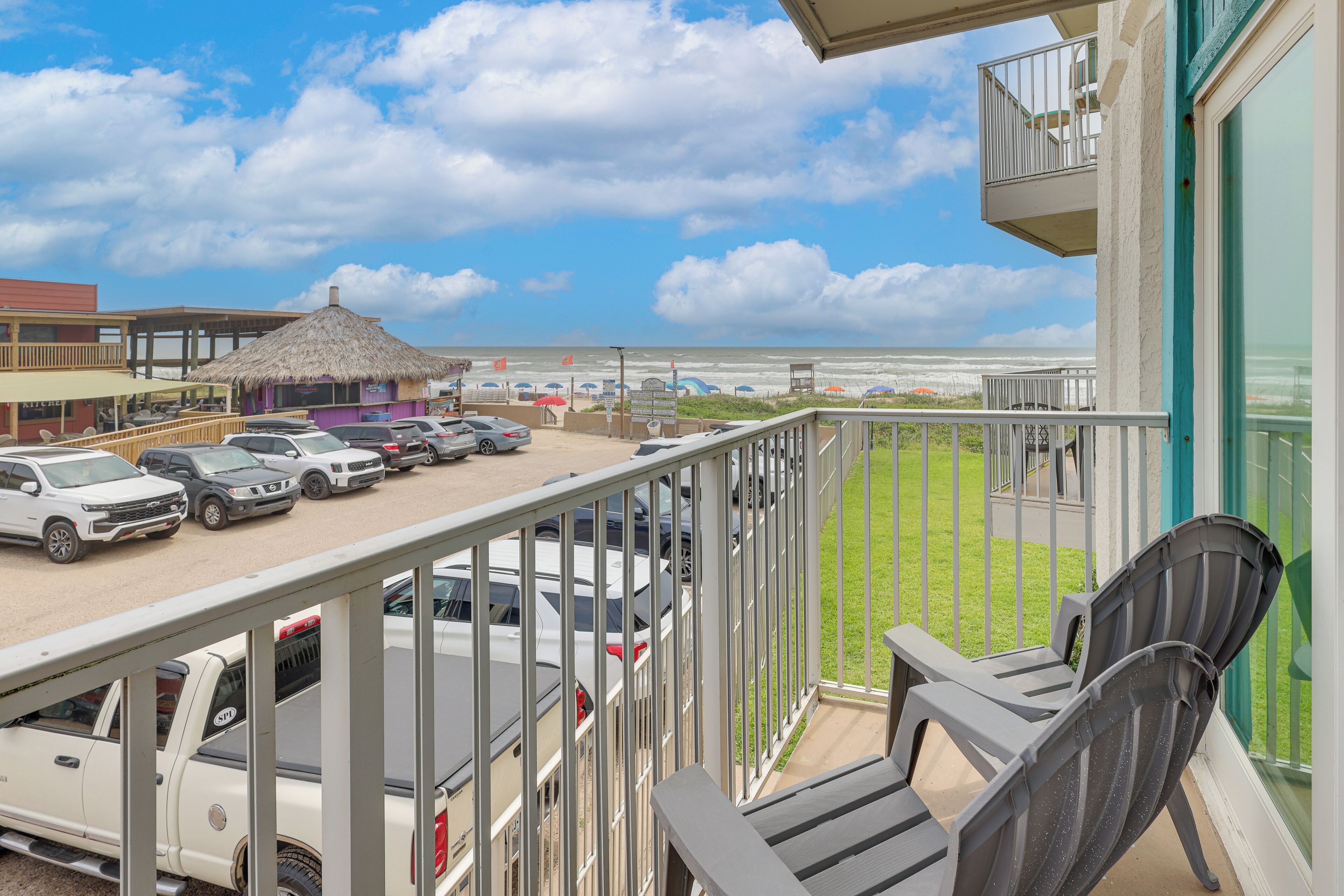 Un balcon avec vue sur la plage, les voitures garées et une construction à toit de chaume.