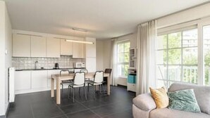 A modern kitchen with white cabinets, a dining area with a table and chairs, and a living area with a sofa and a window with curtains. - 2BR Apartment with Terrace Near Beach (Blankenberge)