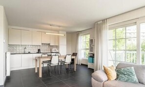Cozinha moderna com armários brancos, área de refeições com mesa e cadeiras, e sala de estar com sofá e janela com cortinas.
