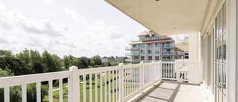 A balcony with a white railing overlooking a green landscape and a multi-story building in the background.