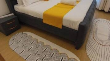 A bed with a dark headboard, white bedding, and a yellow throw. There are two bedside tables, one with drawers and one without. A patterned rug is on the floor, and a circular rug is beside the bed.