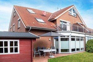 Ein zweigeschossiges Haus mit roter Backsteinfassade, einem Balkon mit Glasgeländer und einer überdachten Terrasse mit Tisch und Stühlen.