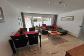 Ein modernes Wohnzimmer mit einer Couch, zwei Sesseln, einem Couchtisch und einem Esstisch mit roter Tischdecke.