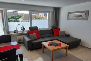 Ein modernes Wohnzimmer mit einer Eckcouch, einem Couchtisch aus Holz und einem Essbereich mit roter Tischdecke.