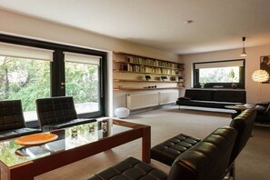 Ein modernes Wohnzimmer mit einem Glastisch, Ledersofas, einem Bücherregal und einem großen Fenster.