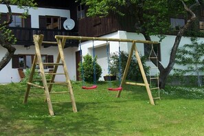 Eine Holzschaukel und eine Kletterleiter in einem grünen Garten mit einem weißen Gebäude im Hintergrund.