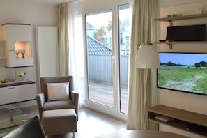 Ein modernes Hotelzimmer mit einer Couch, einem Fernseher, einem Glastisch und einer Schiebetür zum Balkon.