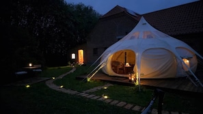 Une tente dotée d’une terrasse en bois, d’une chaise et d’une table à l’intérieur, entourée d’un bâtiment en briques et d’un chemin.