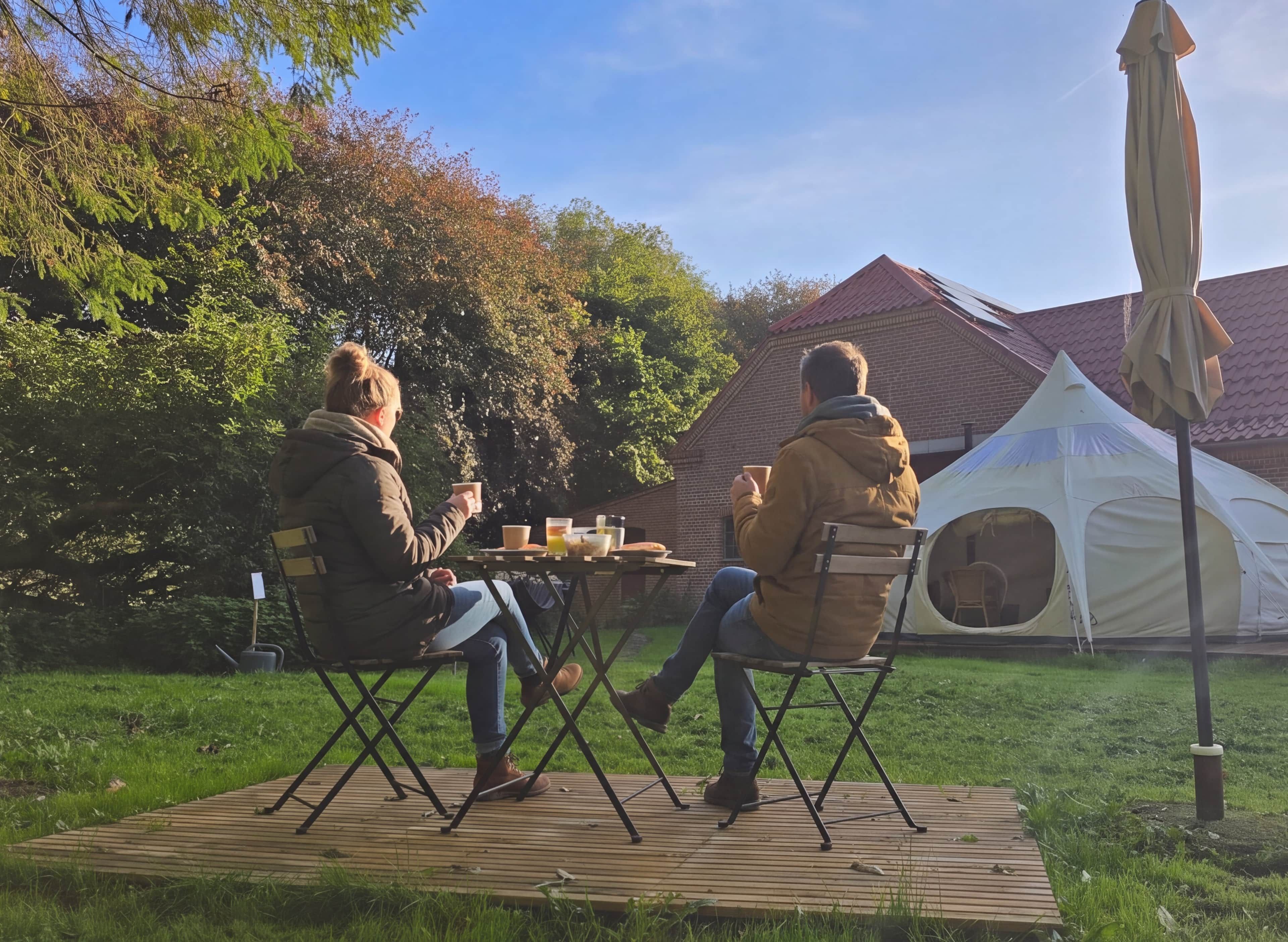 Two people seated at an outdoor table with beverages, a tent, and a grassy area in the background.