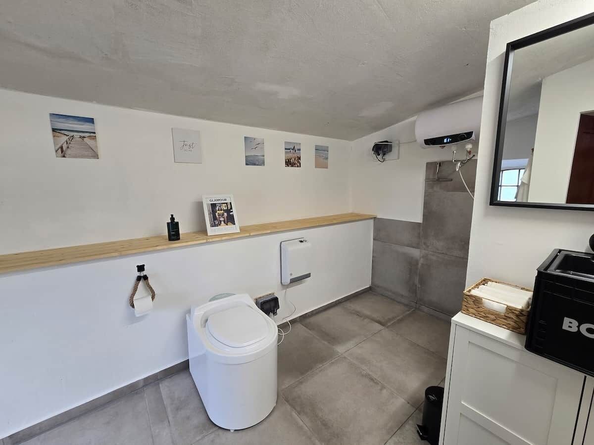 A bathroom with a toilet, sink, and shower. There are pictures on the wall and a small shelf with toiletries.